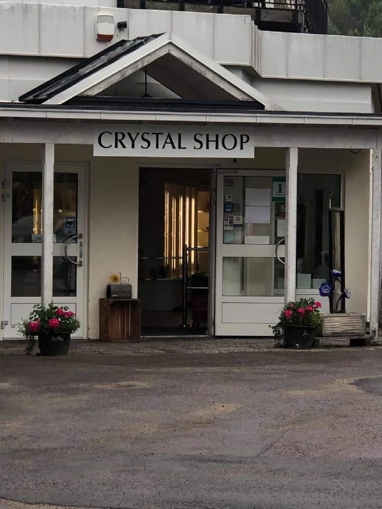 Bild av Målerås Glasbruk, där handgjorda kristallföremål visas i en butiksmiljö. Hyllor i trä visar upp en mängd olika glaskonstverk.