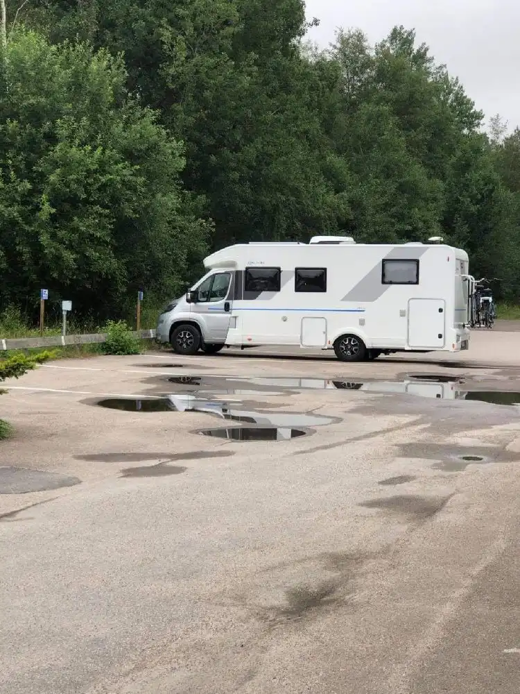 En husbil står parkerad vid Målerås Glasbruk, med en asfalterad parkering och skog i bakgrunden.