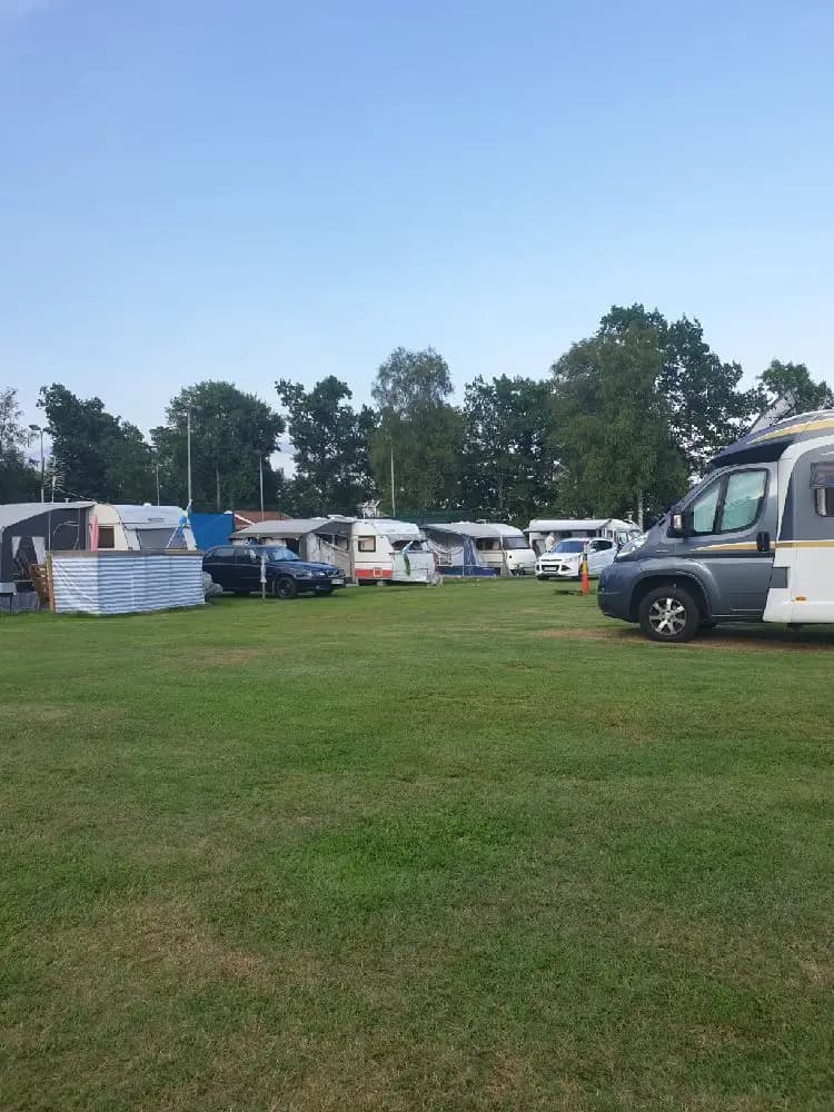 En stor campingplats med flera husbilar och tält uppställda på grön gräsmatta. En parkeringsplats med olika typer av fordon, inklusive lastbilar och husvagnar, syns i bakgrunden.