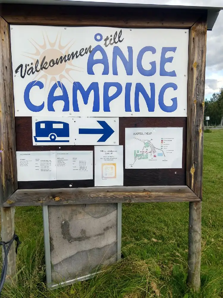 En grönskande campingplats på Ånge Camping med tält, husvagnar och en bil intill stora träd. I bakgrunden syns en husvagn i parken och en lastbil med presenning. En lugn och naturnära miljö för utomhusliv.