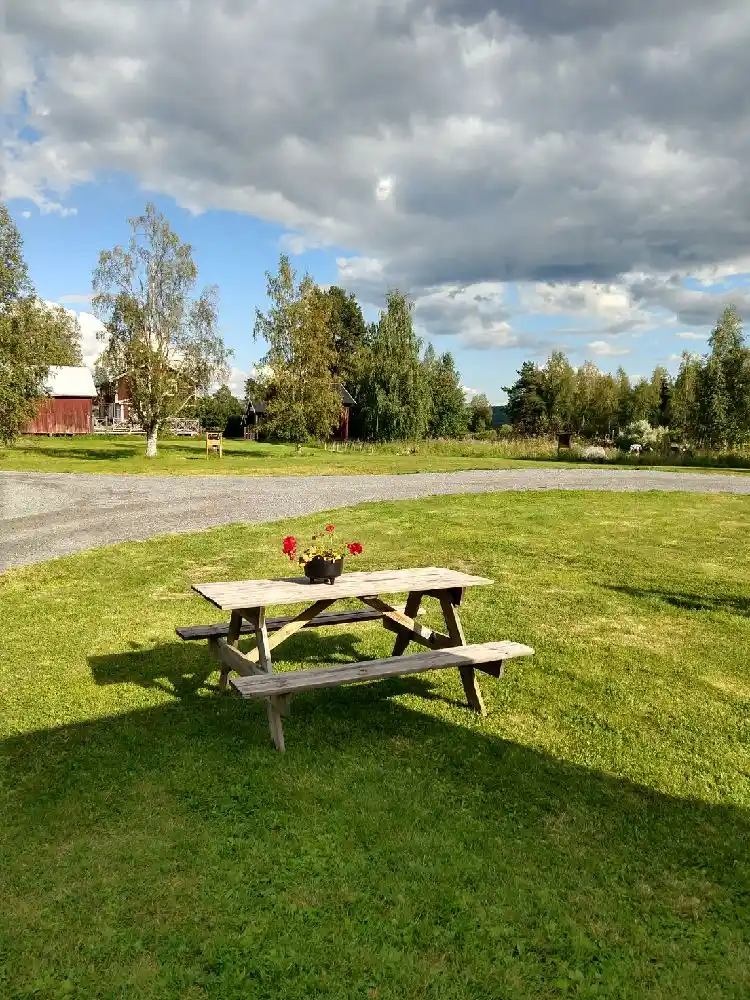 En grön gräsmatta under en blå himmel med moln på Ånge camping. I förgrunden syns en picknickbänk och utemöbler placerade på det stora öppna fältet.