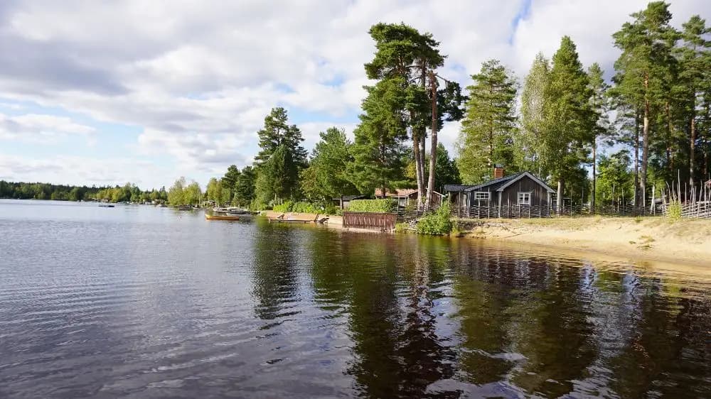 En sjö omgiven av träd och stugor vid Högbacka camping, med en stilla vattenspegel och skogsbeklädda kullar i bakgrunden.