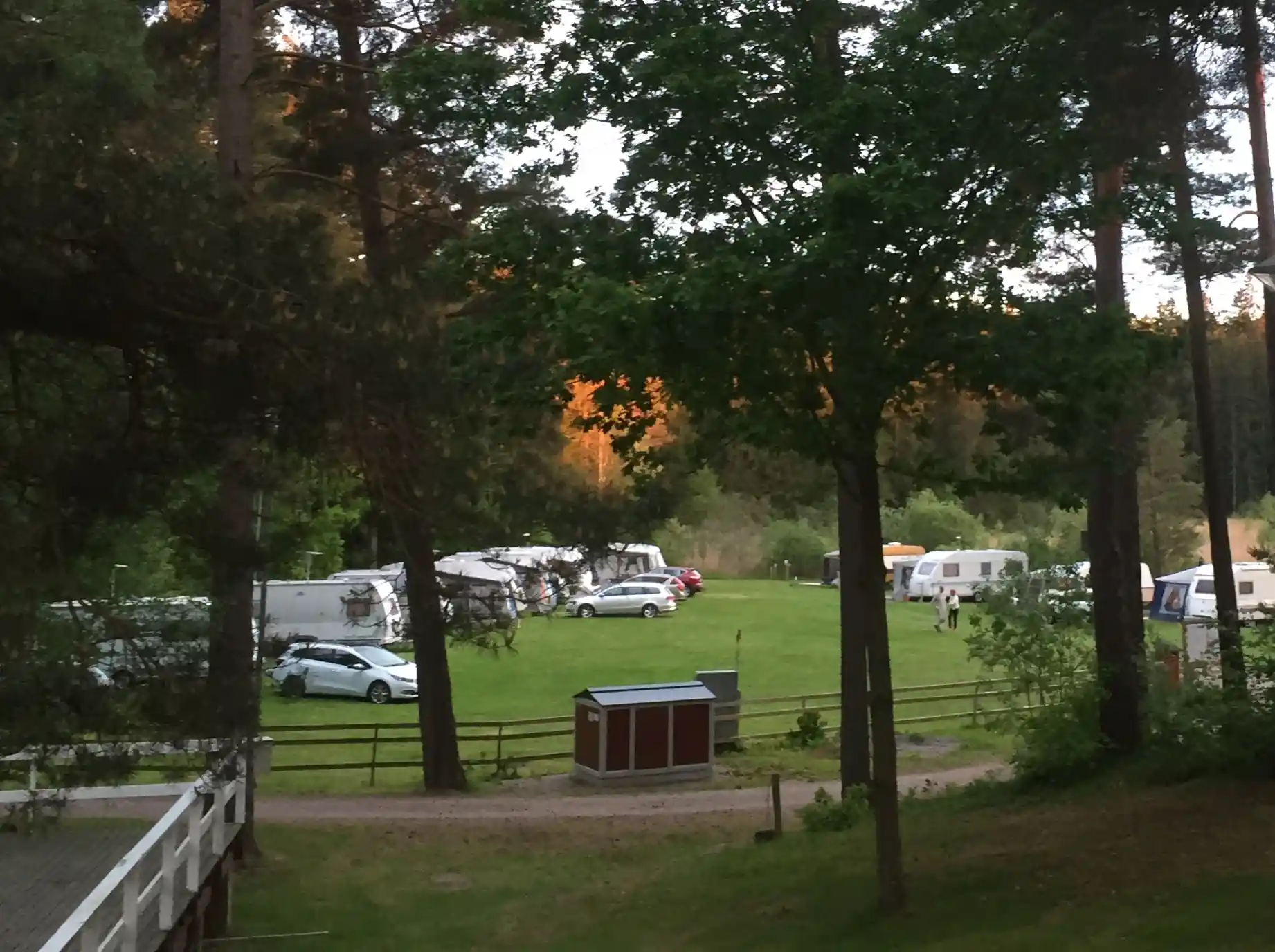 En campingplats med parkerade bilar i närheten av en skogssjö. Träden omger området och det finns en gräsyta intill parkeringsplatsen. Olika typer av fordon, inklusive familjebilar och husbilar, syns i bild.