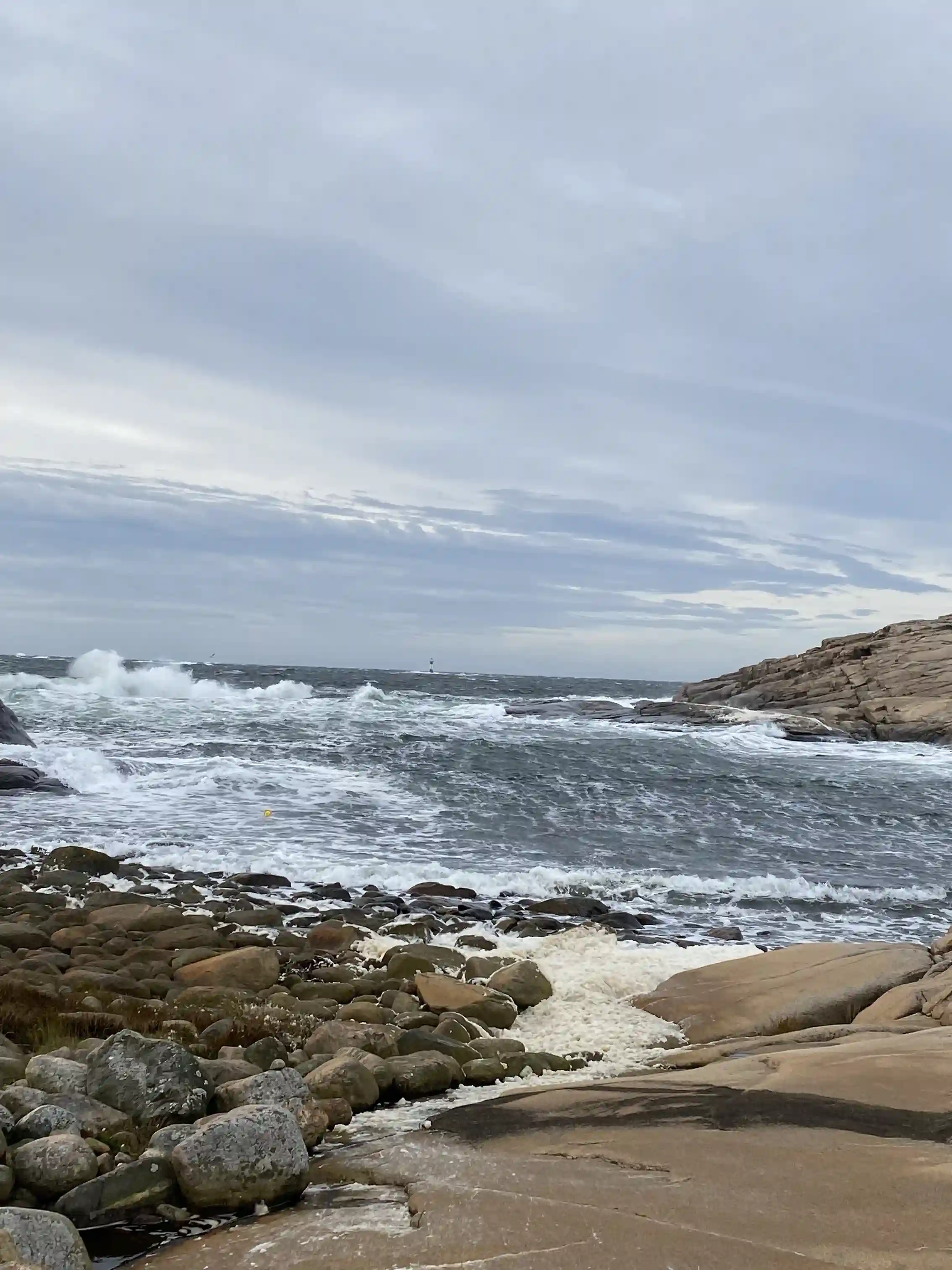 Vy över kustlandskap vid Saltviks camping med klippor och havet i förgrunden. Det är en molnig dag med vågor som slår mot stranden och ett synligt horisontlinje i bakgrunden.