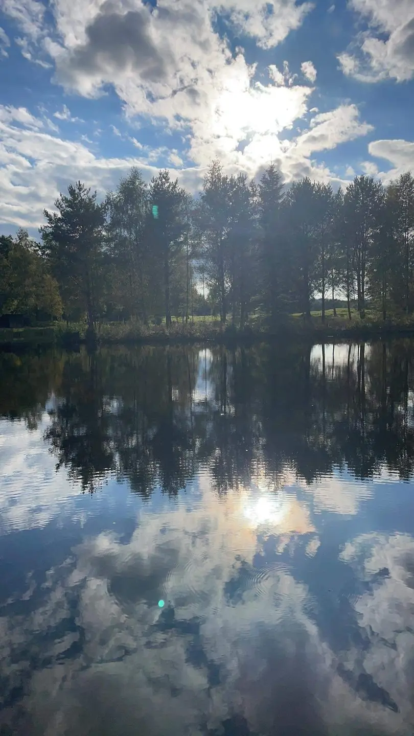 En klarblå himmel speglas i det stilla vattnet vid Våxtorps naturcamping, omgiven av grönskande natur och mjuka moln.