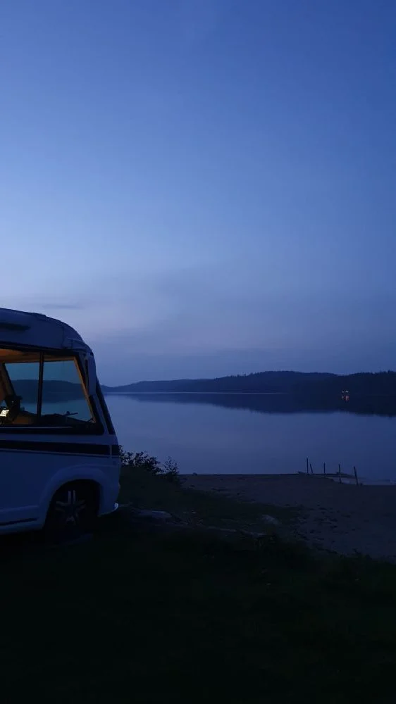 En husvagn står parkerad intill en sjö i skymningen, med en vacker solnedgång i bakgrunden.