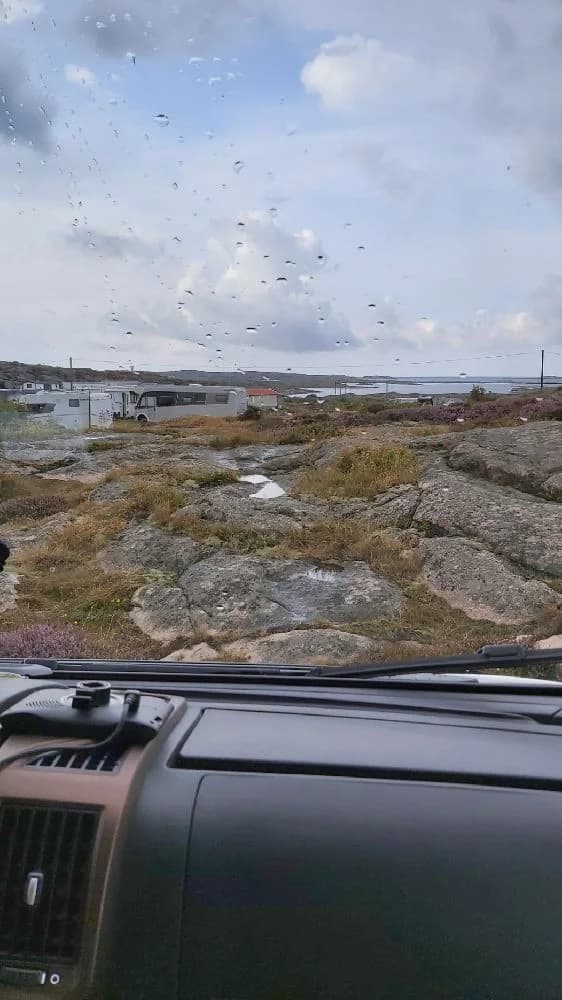 En bil kör längs en väg genom tundralandskap, med moln på himlen. Vindrutetorkare och bilspeglar syns i förgrunden.