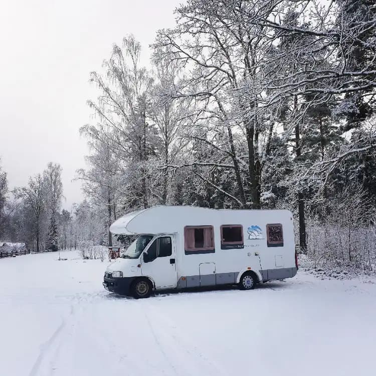 En vintrig campingplats med snö och frost täcker marken. Flera husbilar och skåpbilar står parkerade, omgivna av snötäckta träd. Himlen är grå och det är tydligt att det nyligen snöat.