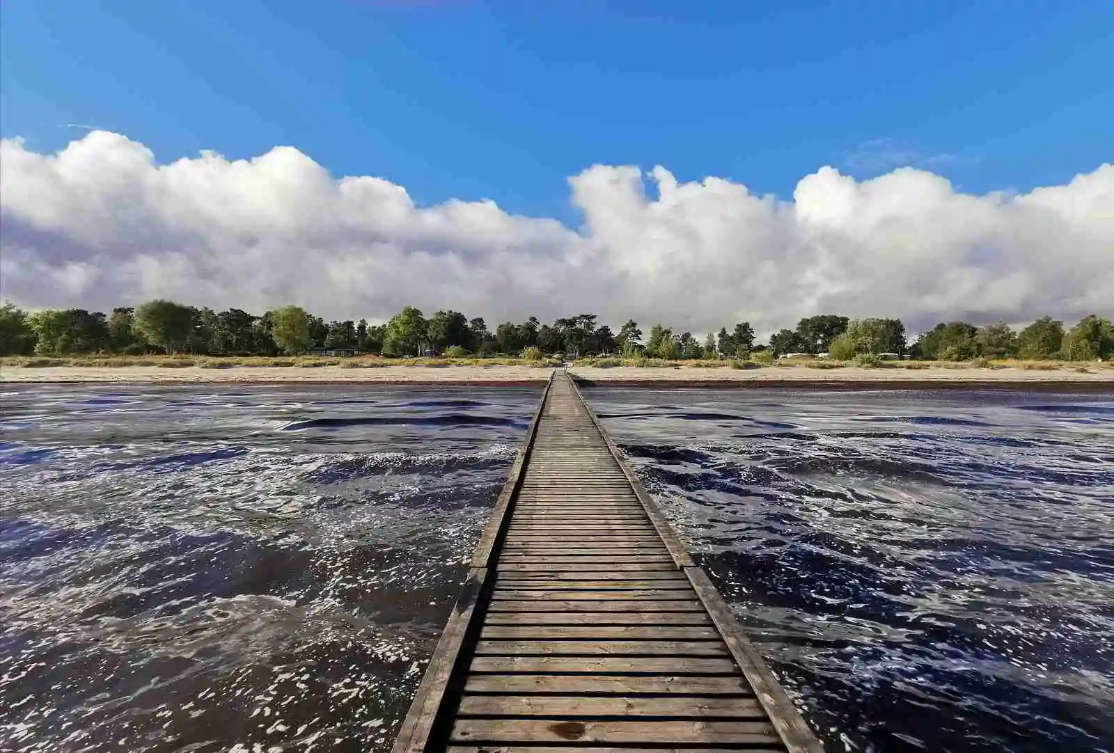 Vy över kusten vid Bingsmarkens camping med en molnig himmel i horisonten. En brygga sträcker sig ut över havet, vilket ansluter till stranden och skapar en idyllisk kustmiljö.