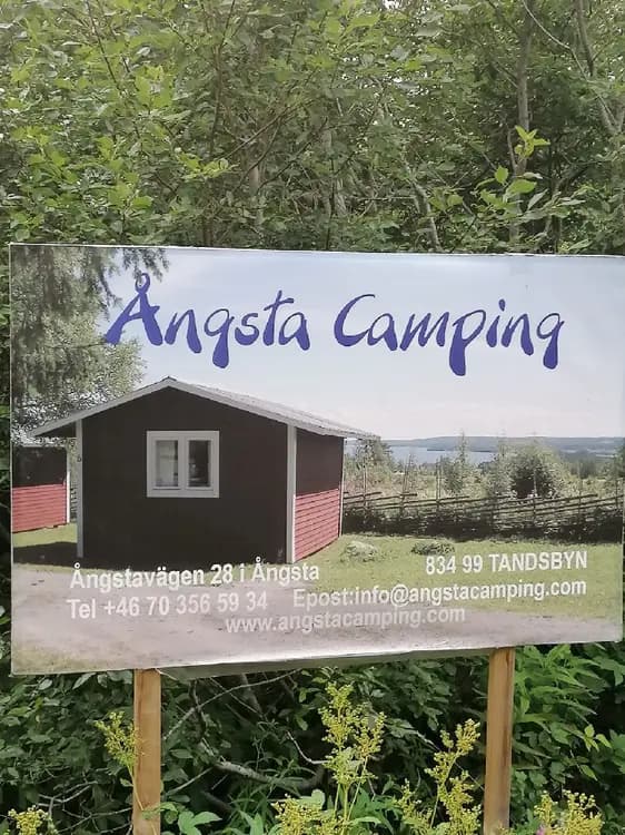 En grönskande campingplats med en skylt som välkomnar besökare till Ångsta Camping. På området finns trädgårdsbyggnader omgivna av välskötta gräsmattor.