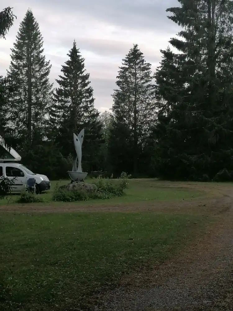 En frodig campingplats med grönskande träd och välskötta gräsmattor, omgiven av barrträd som granar och tallar.