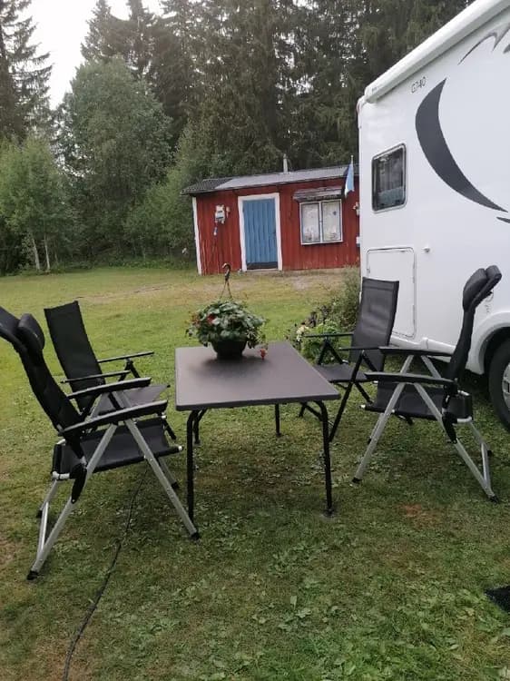 En campingplats med utemöbler, inklusive stolar och ett bord, belägen i en grönskande trädgårdsmiljö med gräsmatta.