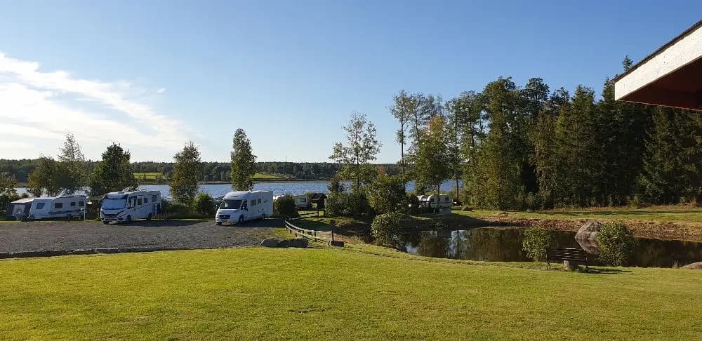 En campingplats med husbilar och husvagnar parkerade nära en sjö. Omgiven av grönskande natur, med vatten som glittrar i bakgrunden.