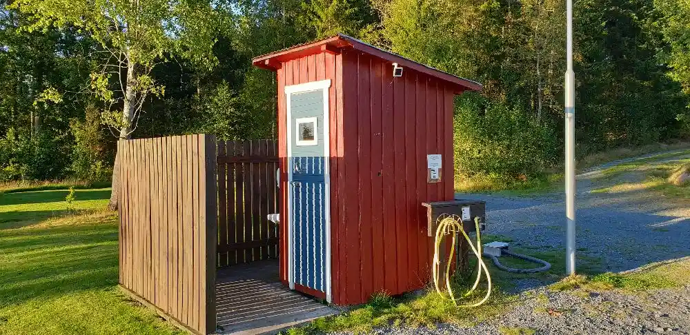 En liten träbyggnad med brun träpanel står bland grönska, omgiven av utemiljö typisk för en campingplats.