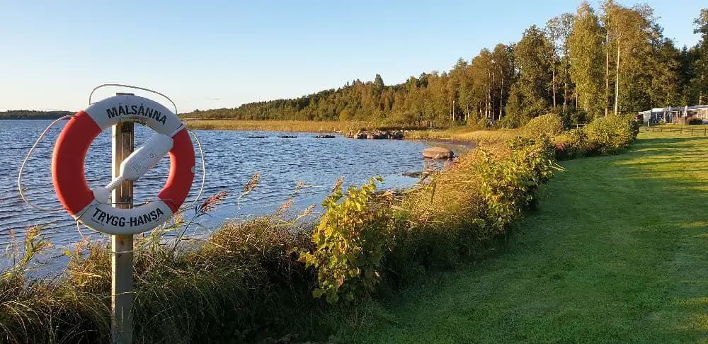 Vy över Målsånna Camping Park, som visar en sjö med klart vatten och omgivande stränder. I bakgrunden syns skogsbeklädda landformer, vilket skapar en avkopplande och naturnära miljö för besökare som söker rekreation vid vattnet.