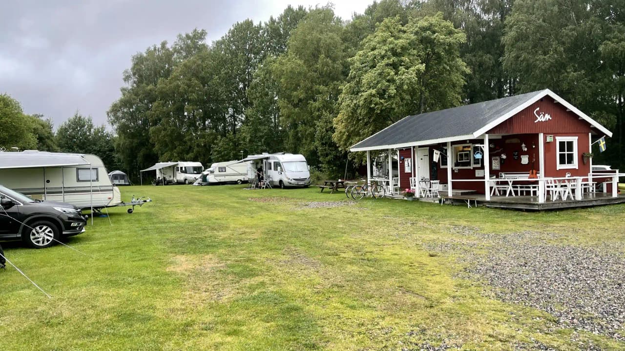 En somrig campingplats med husvagnar och husbil uppställda på gräsytor, omgiven av gröna ängar. I förgrunden syns en stuga med caféverksamhet och parkeringsytor, allt beläget i en lantlig och naturskön miljö.