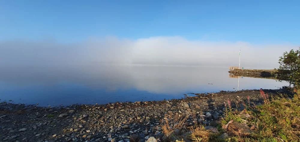 En vy över Jormvattnets Fiskecamp med en klarblå sjö i förgrunden och ett vidsträckt horisont i bakgrunden, omgiven av klippformationer och ett öppet kustlandskap.