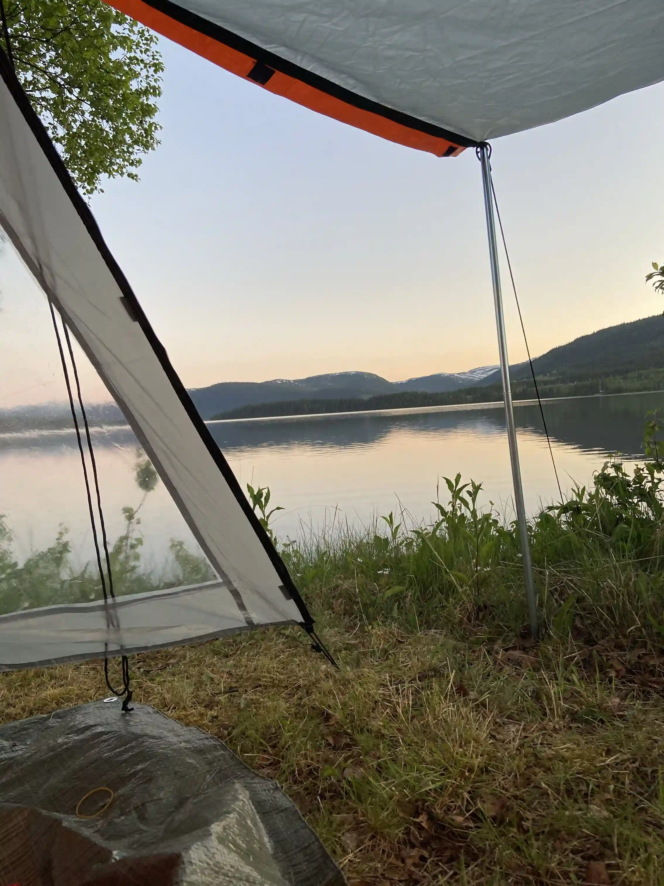 En tältplats vid Jormvattnets fiskecamp med vy över en stilla sjö omgiven av lummig natur. Tältet står på en grön gräsmatta med en sjö i bakgrunden och omgärdas av skuggande träd.