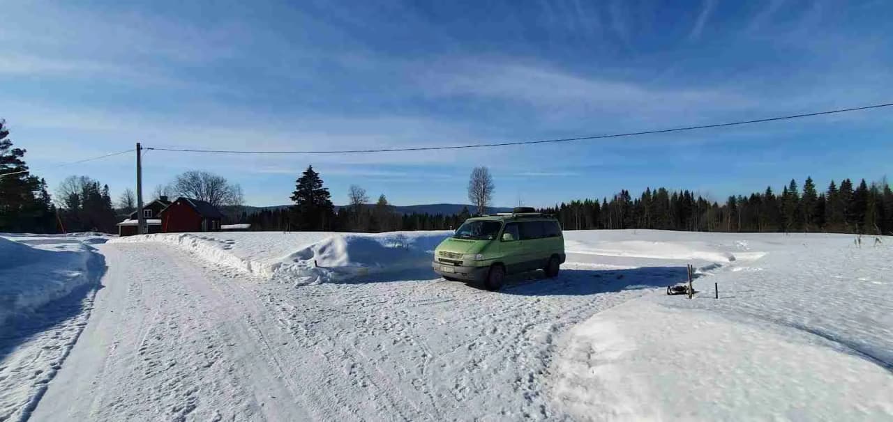 En snötäckt vinterlandskap med en bil parkerad vid vägkanten. Skåpbilen, täckt av frost, är delvis synlig med fokus på sidospegeln och bildörren.
