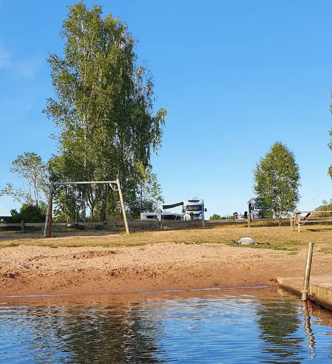 En husbil parkerad vid en ställplats intill en skogskant med utsikt över en lugn sjö, omringad av frodig natur och våtmarker.