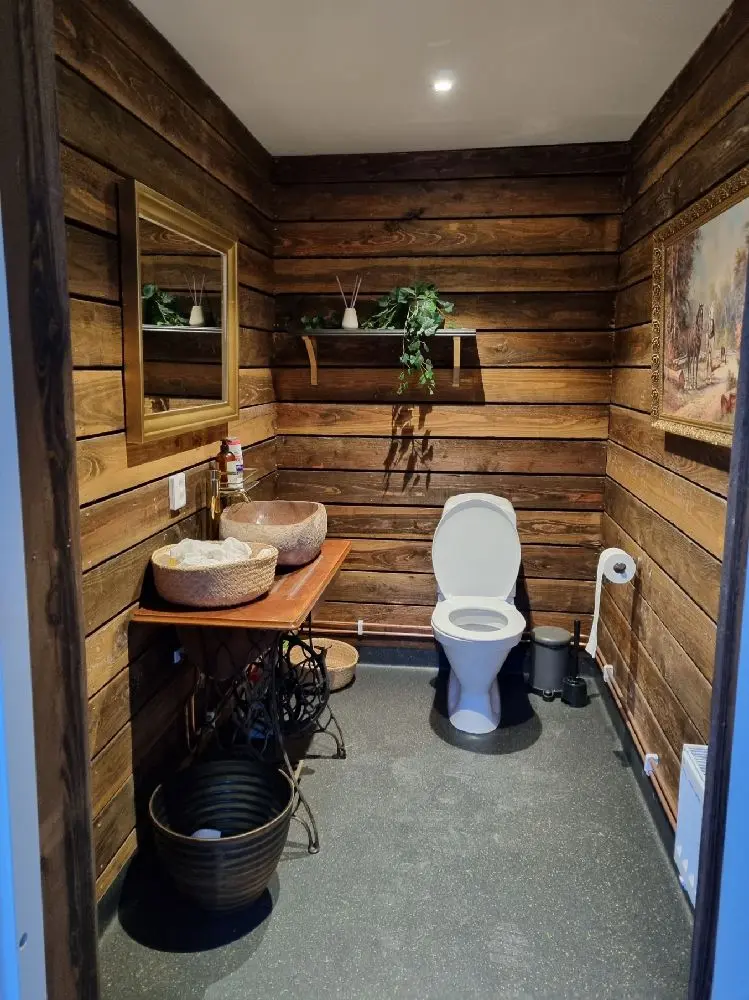 En enkel och rustik badrumsinteriör med träbetonade detaljer, inklusive ett trätoalettlock och trägolv, visas.