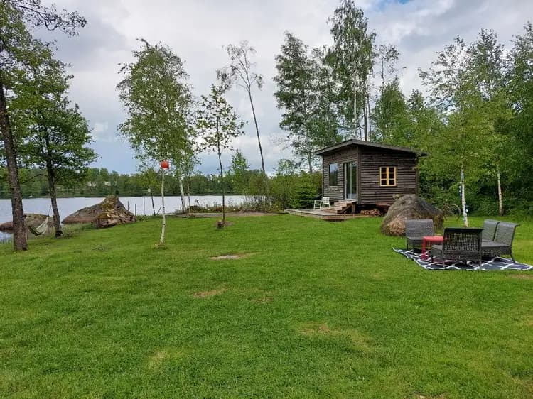 Ett lantligt område med en stuga omgiven av en välskött trädgård, där finns även en gräsmatta, ett skjul och olika växter.