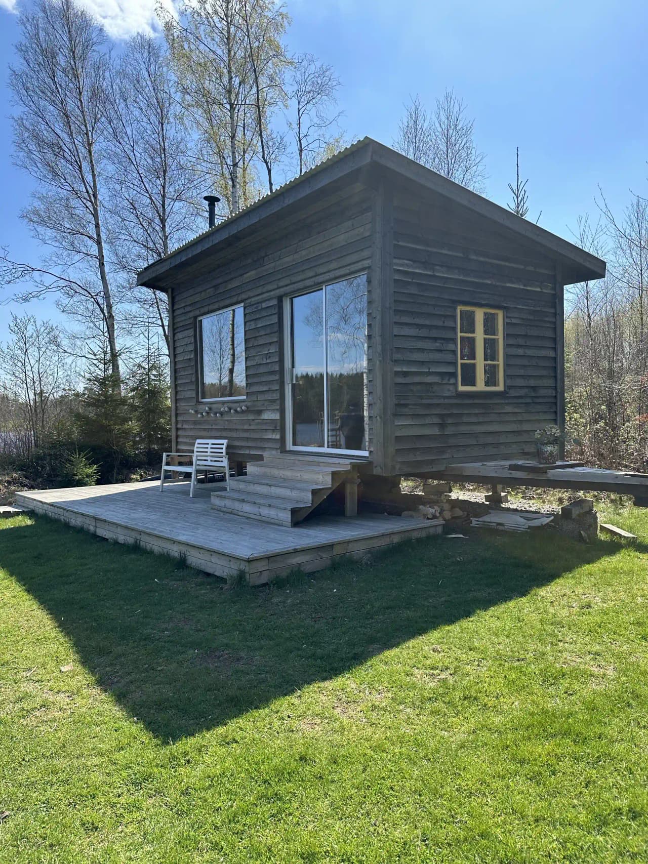 En trästuga med stående träpanel och en enkel dörr, omgiven av grönska.