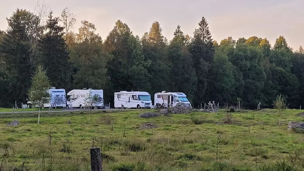 En husvagn och en husbil står parkerade på en campingplats omgivna av träd. I bakgrunden syns tält och en lastbil, vilket skapar en avslappnad atmosfär för besökare på Stjernkvist's Hunshult.