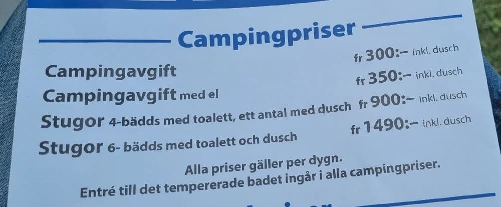 Bild av Töreboda Camping & Bad som visar ett campingområde med tält och husvagnar uppställda i en grönskande parkmiljö. Skuggor från trädtält och markiser ger en känsla av avkoppling och semester.