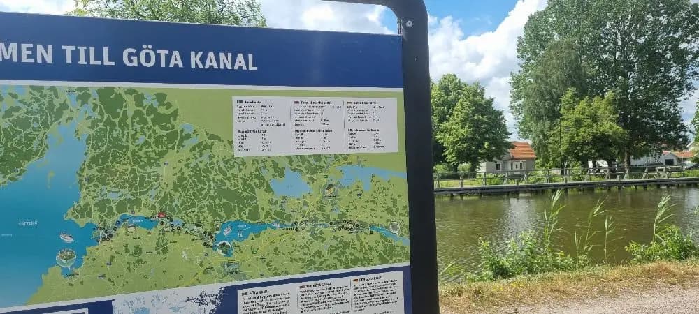 Bild av Töreboda Camping & Bad, med en skylt i förgrunden som visar platsens namn. I bakgrunden syns ett grönområde med en liten damm omgiven av träd och öppna ytor, vilket ger en naturskön miljö.