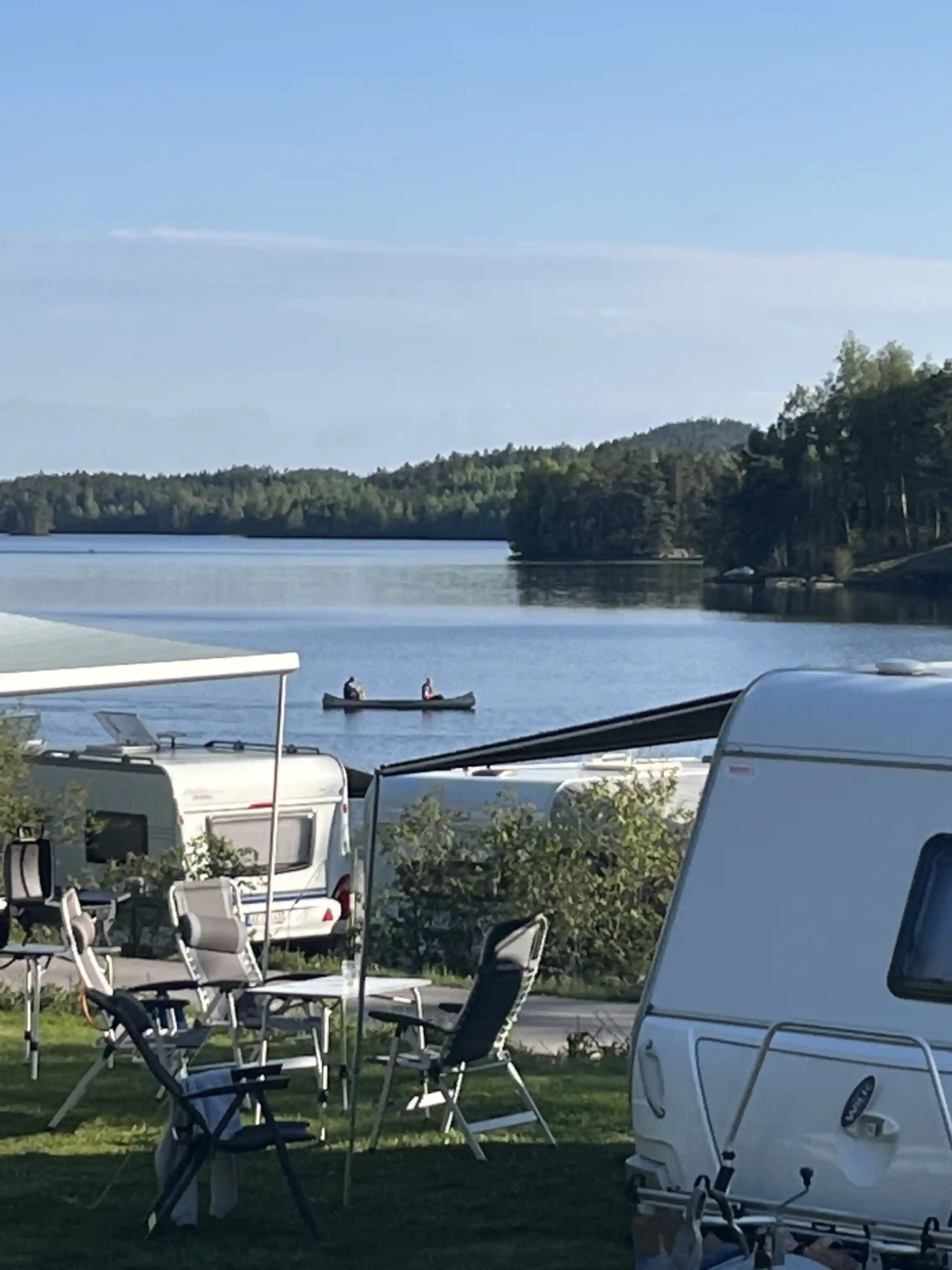 En husvagn parkerad vid Ragnerudssjöns camping och stugby, med en sjö i bakgrunden där en båt ligger vid en brygga. Campingområdet är omgivet av natursköna omgivningar med utemöbler vid vattnet.