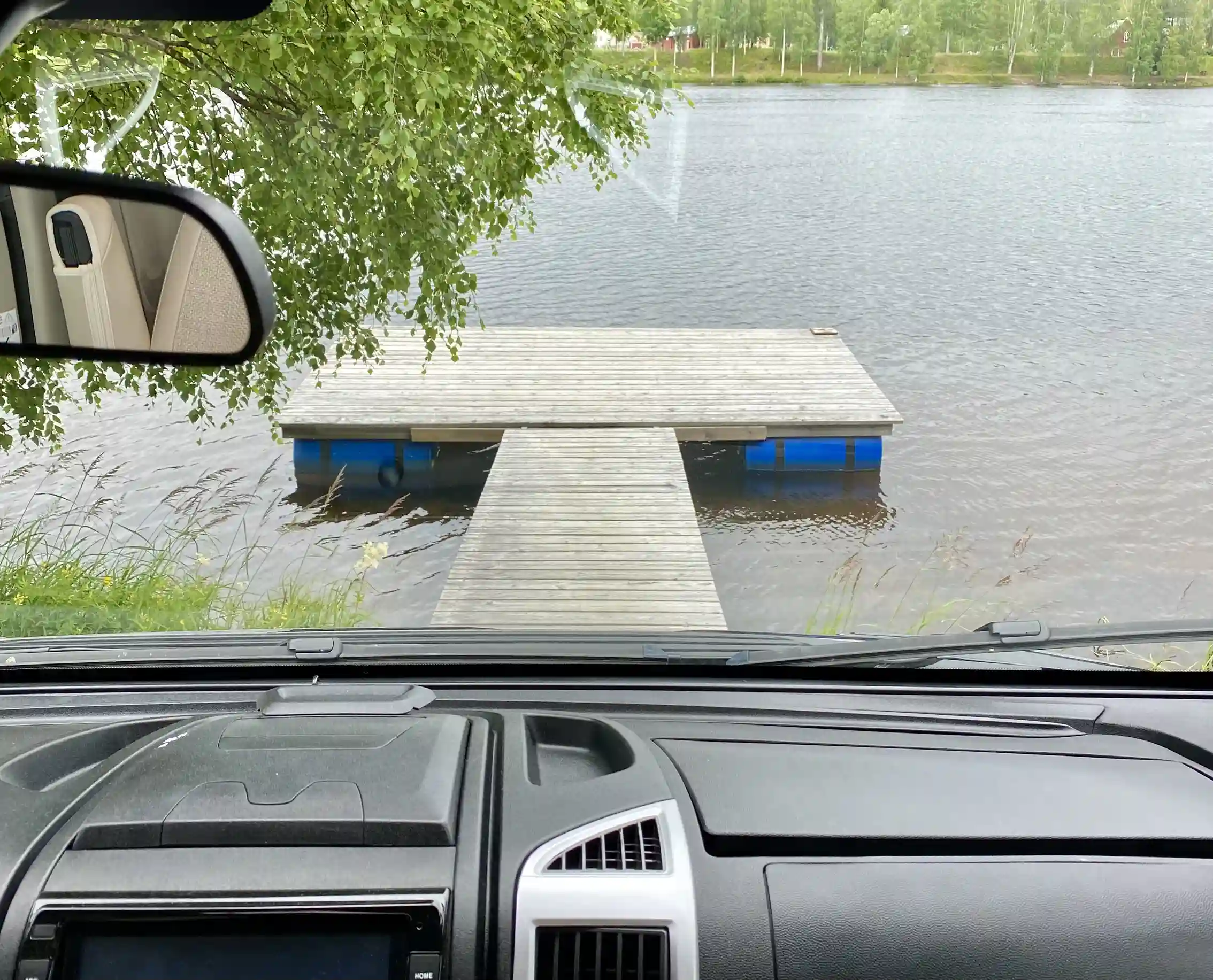 Vy av en stilla sjö med spegelblanka ytor som reflekterar himlen, sedd genom bilfönster. I förgrunden syns delar av en bilruta med backspegel och vindrutetorkare, vilket ger en känsla av att vara på resande fot vid besöksmålet Granö Beckasin.