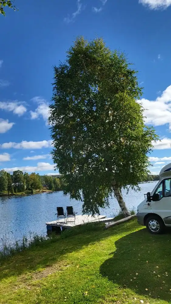 En naturskön miljö vid Granö Beckasin med en lugn flod som slingrar sig genom landskapet, omgiven av frodigt gräs och lummiga träd. En stilla sjö speglar omgivningarna under en klar himmel.