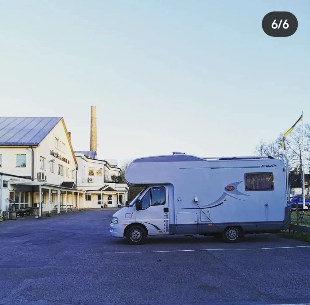 En parkeringsplats vid Målerås Glasbruk med flera olika typer av fordon, inklusive husbilar och skåpbilar, som är parkerade i soligt väder.