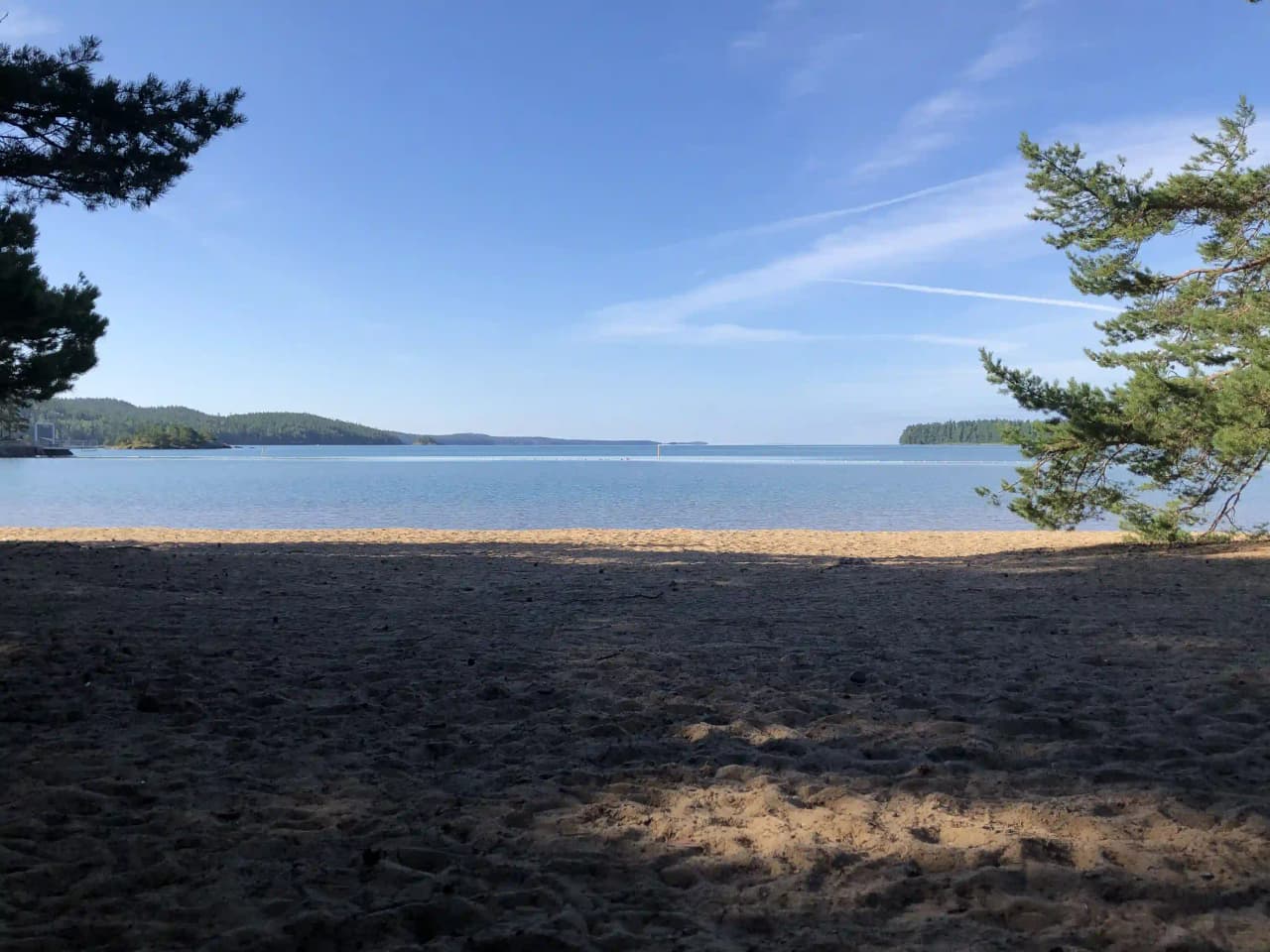 En solig dag vid Harge Bad & Camping med klarblå himmel, glittrande vatten och en sandstrand omgiven av naturlig grönska.