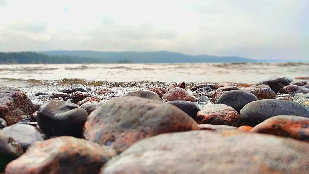En bild av Harge Bad & Camping som visar en klippig kustlinje med stenar och småstenar vid stranden. I bakgrunden syns havet under en kvällshimmel.