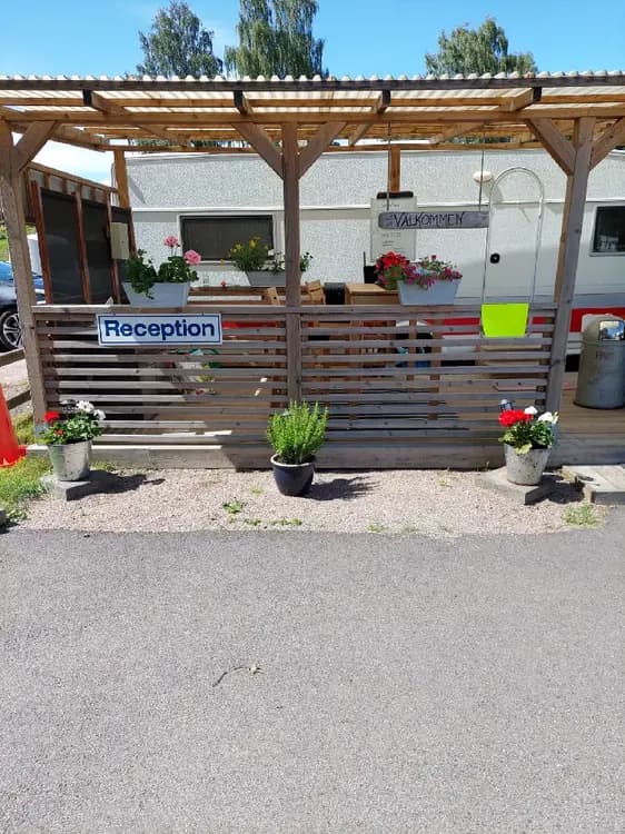 En solig dag på Öjaby Camping med en pergola som ger skugga över uteplatsen. Blomkrukor pryder området och trädgårdsmöbler står redo på gräsmattan. En staket omger den lugna platsen som bjuder in till avkoppling.