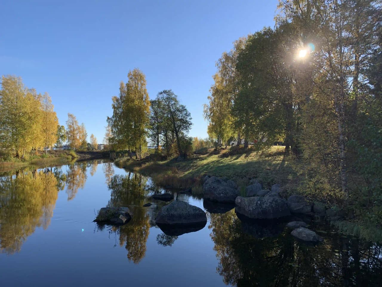 En naturskön vy vid Ljusnefors camping med ett lugnt vattendrag som speglas av omgivande grönska och stenar längs strandkanten.