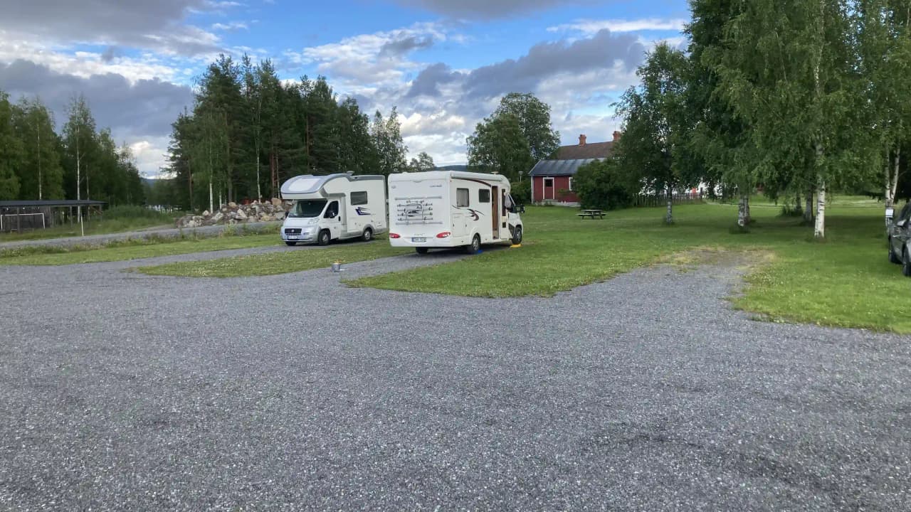 En campingplats med flera parkerade husbilar och husvagnar på ett grusbelagt område, omgiven av gröna träd och gräsmattor. Perfekt för uppställning och avkoppling i naturskön miljö.