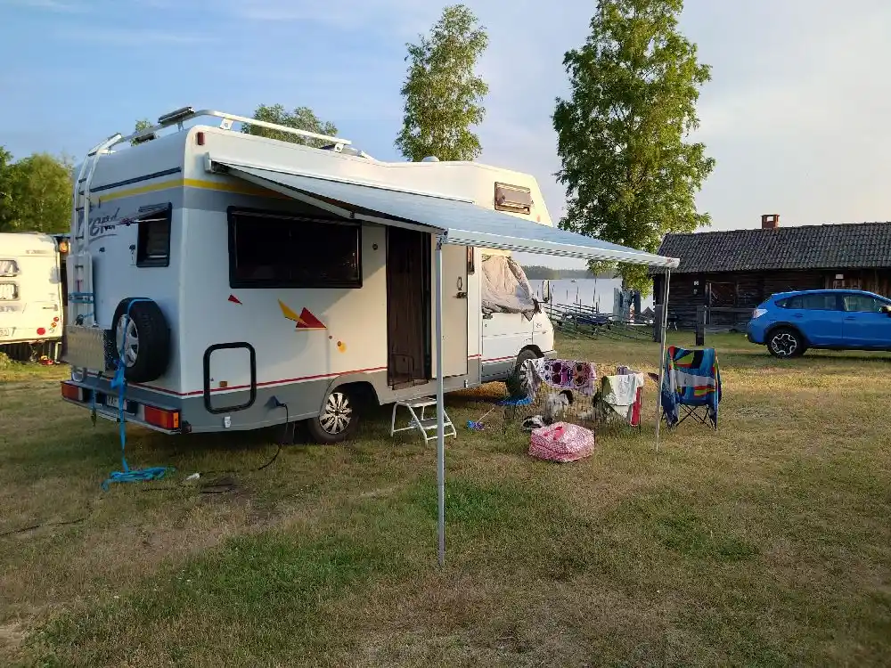 En campingplats med flera husbilar och husvagnar uppställda, omgivna av grönska. Flera fordon syns, inklusive vans och mindre lastbilar, med campingutrustning lastad.