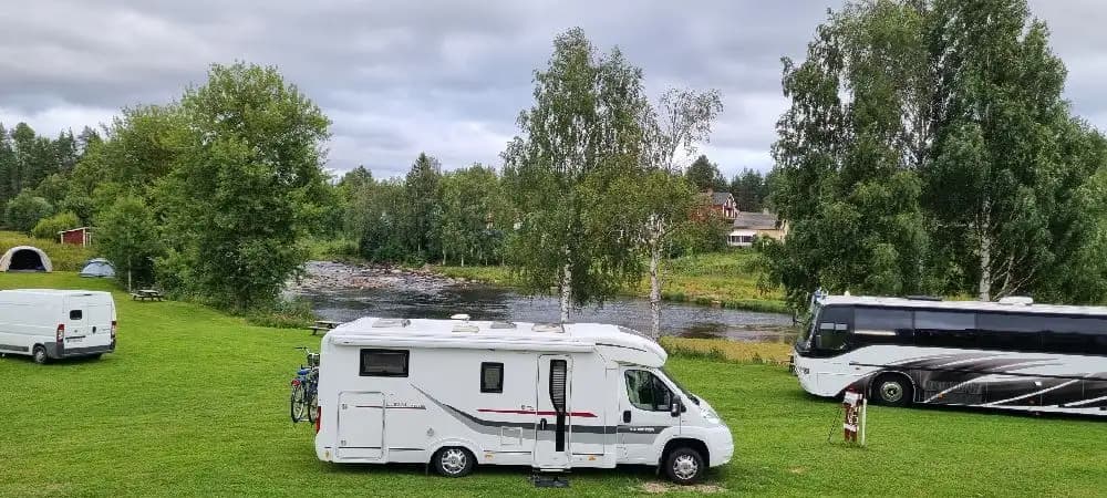 Alt-text: En husbil och en husvagn parkerade vid Åmsele camping, omgivna av grönska och med andra campingfordon i bakgrunden.