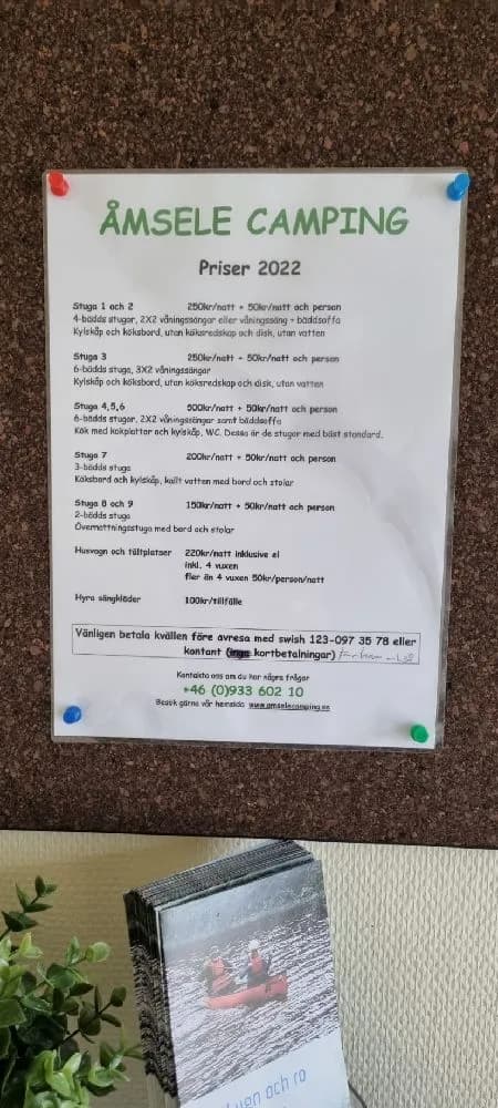 En skylt vid infarten till Åmsele camping, omgiven av naturskön miljö med träd och grönska. Skylten annonserar campingfaciliteter och lockar besökare till platsen.