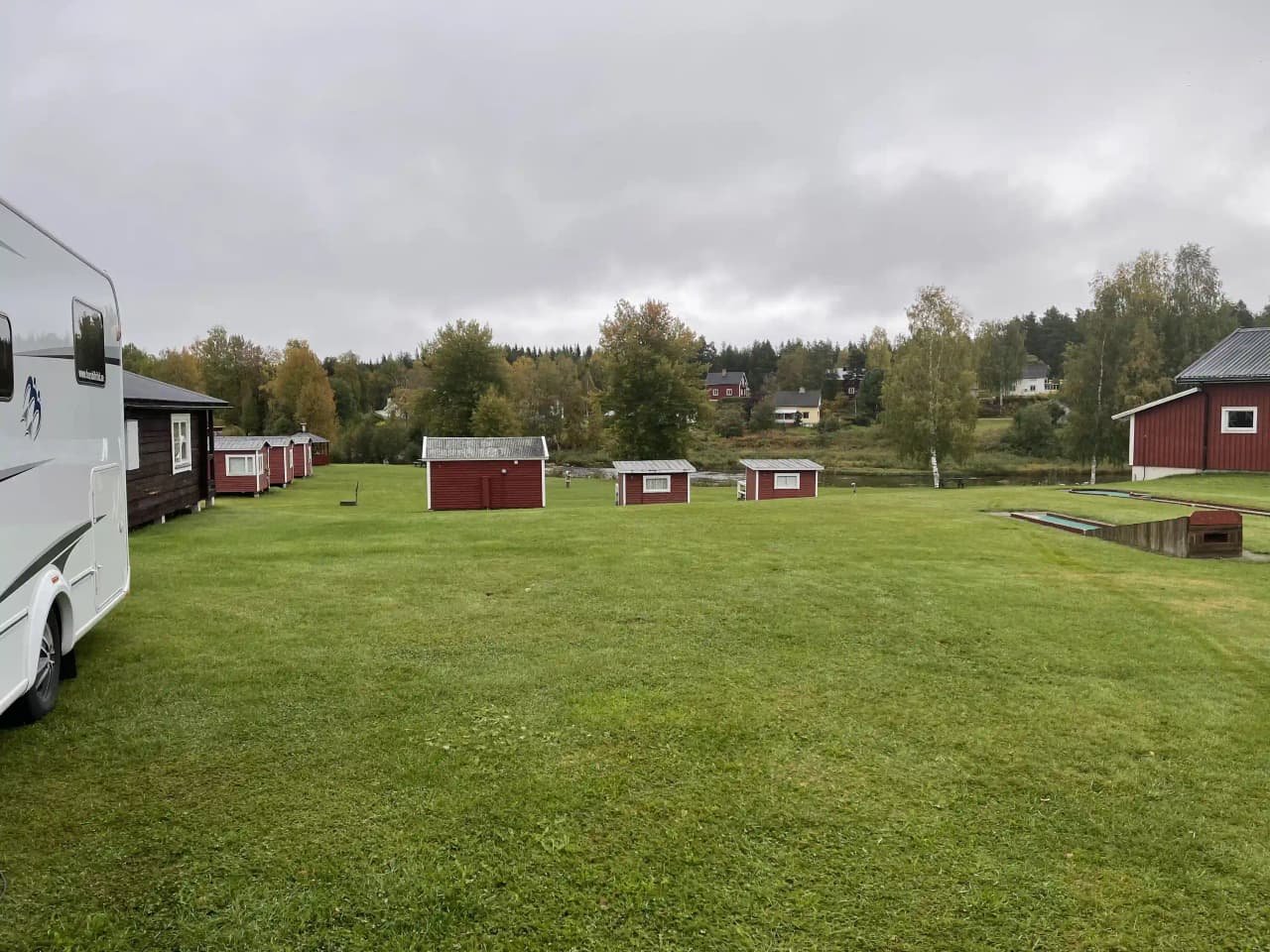 En grönskande campingplats med stora gräsytor och utspridda träd. På bilden syns en öppen äng omgivet av trädgårdsbyggnader och välskött landskap, vilket skapar en inbjudande miljö för besökare.