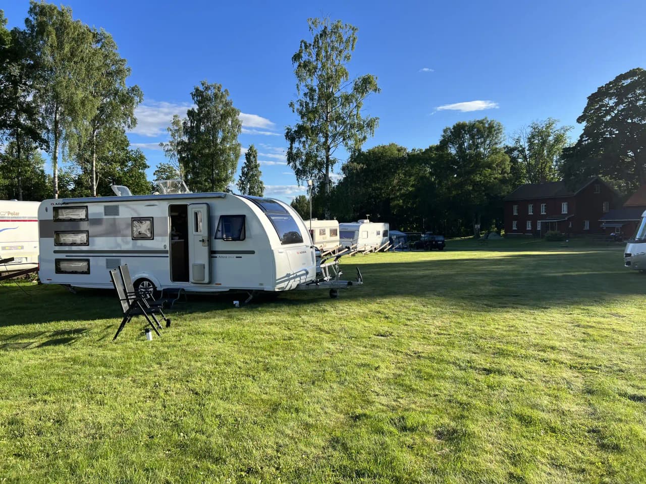 Alt-text: "En campingplats vid Furusjön med flera husvagnar och husbilar parkerade på en gräsbevuxen yta, omgiven av natur."