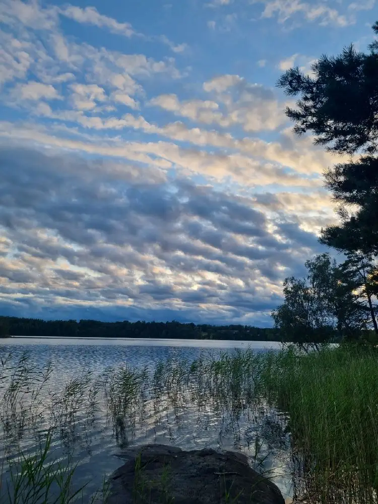 Bild av Furusjöns camping med en klar blå himmel och moln i bakgrunden, speglat i det lugna sjövattnet. En naturskön plats vid vattnet.