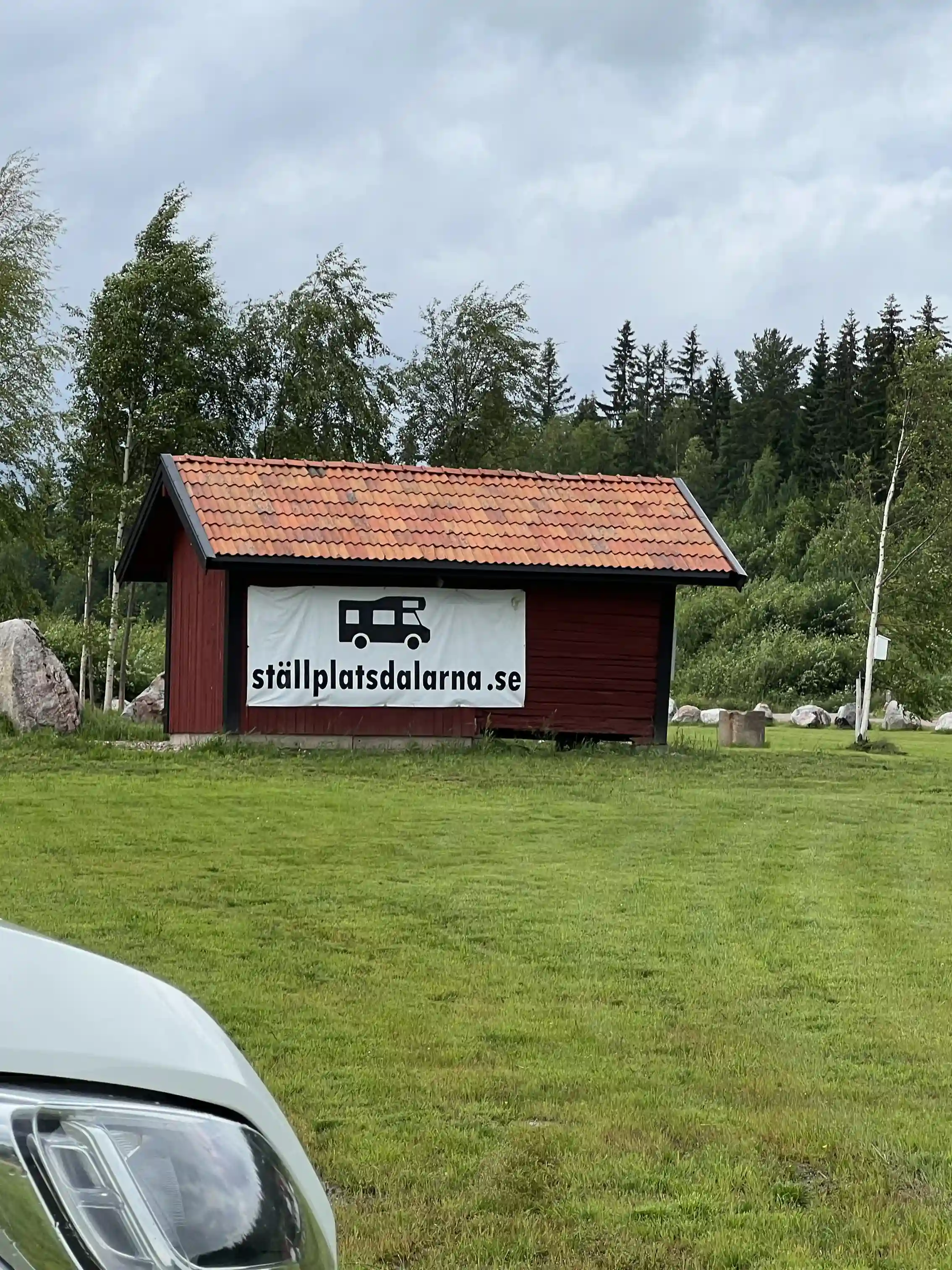 En ställplats i Dalarna med en grusad uppfart, ett litet skjul och skyltar i närheten, omgiven av grönska.