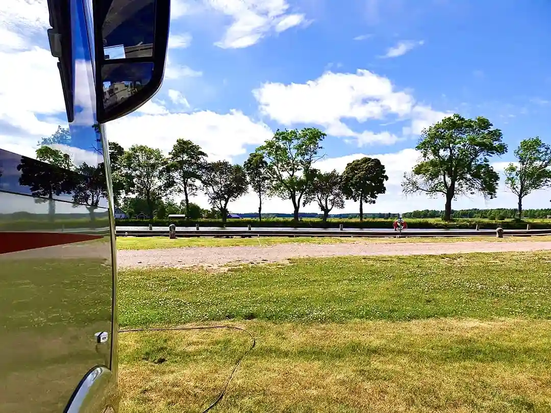 En turkos turistbuss står parkerad på en grön gräsmatta under en molnig himmel. En sidospegel på bussen är synlig, och i bakgrunden breder ett öppet landskap ut sig.