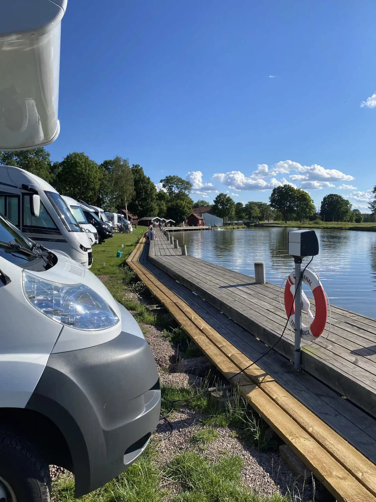 En gångbrygga sträcker sig över vattnet i en park, med rekreationsfordon i bakgrunden vid Jonsboda.
