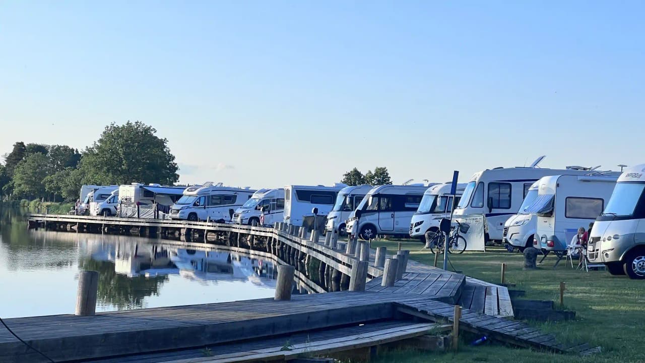 En husbil och en husvagn parkerade nära en kanal vid en småbåtshamn, med bryggor och båtar reflekterade i vattnet.