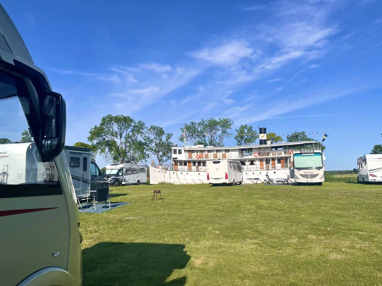 En husvagn parkerad på en grön gräsmatta i skuggan av träd, med en lastbil bredvid inom en parkmiljö.